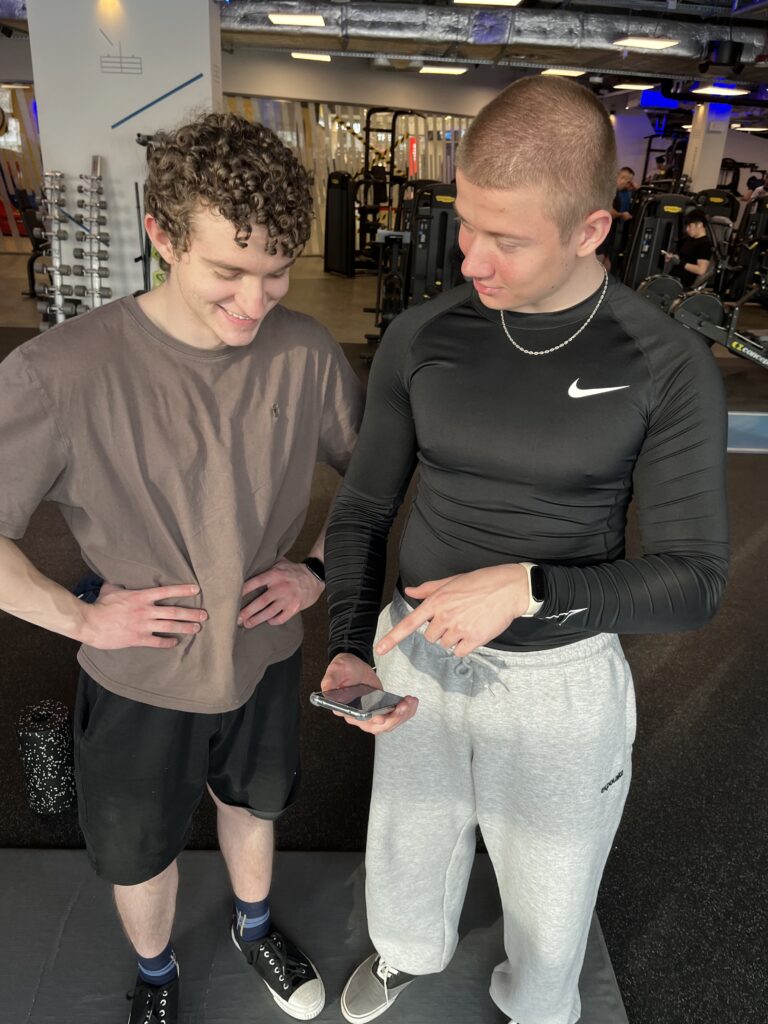 Trener personalny CityFit PDT Wola Adrian Wyszomirski omawia z podopiecznym na smartfonie dane dotyczące optymalizacji regeneracji treningowej na siłowni.